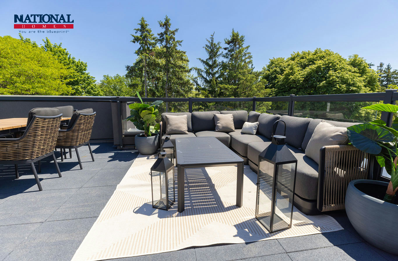 Rooftop patio furnished with a grey outdoor sectional, wicker dining chairs, lanterns and potted plants, with tall trees beyond