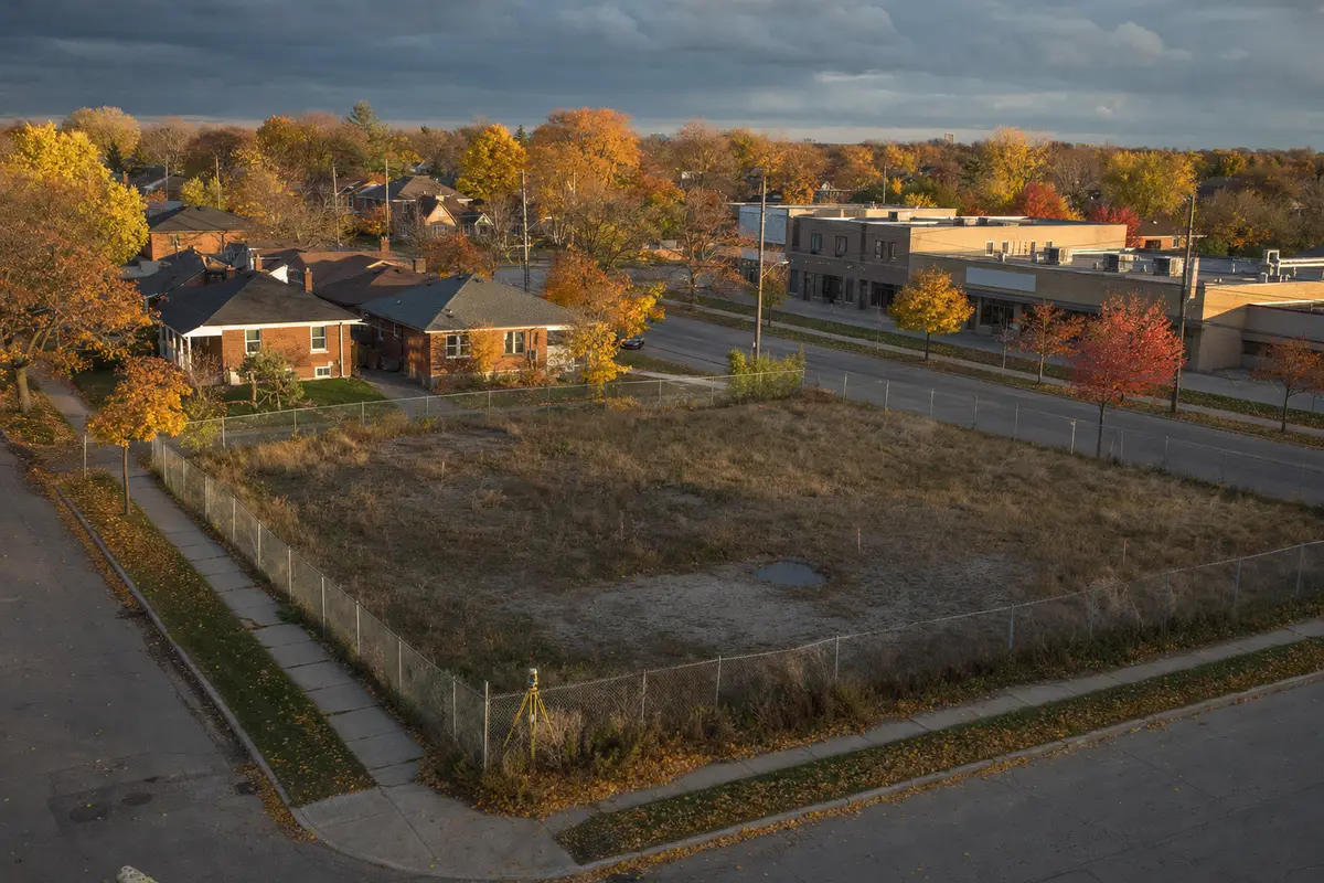 Fenced vacant land parcel with survey stakes beside an older brick neighbourhood in autumn